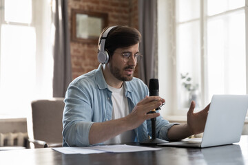Millennial man streamer wear headphones take part in virtual communication, records podcast seated at desk, speaking reflections on topic, make speech into microphone, create audio file. Tech concept
