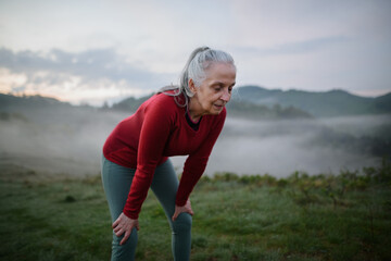 Obraz premium Senior woman doing breathing exercise in nature on early morning with fog and mountains in background.