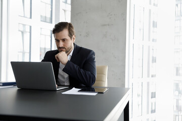 Serious focused businessman working use laptop seated at modern skyscraper office, solve business...