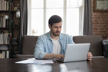 Fototapeta premium Serious office employee sit at desk working on laptop, prepare report, makes on-line presentation looks focused at homeoffice workplace. Workflow, professional business application, tech usage concept