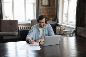 Man in headset writing on copybook while listen on-line tutor during distancing class via video...