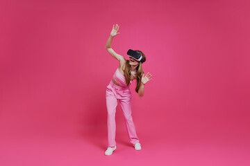 Full length of cheerful young woman in virtual reality headset having fun against pink background