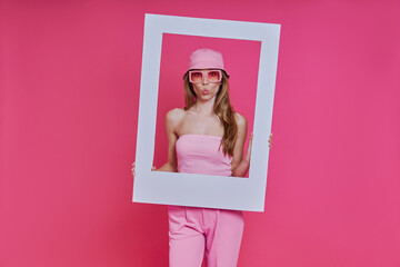 Playful young woman looking through a picture frame while standing against pink background