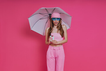 Attractive young woman in trendy clothing holding umbrella while standing against pink background