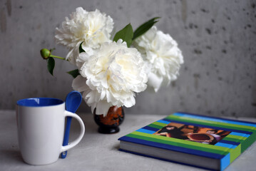 On a table is a bouquet of white peonies in a vase, next to it is an book, a glass of coffee or tea. Bokeh background.