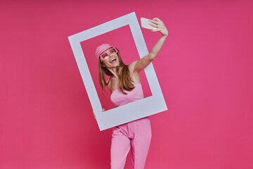 Playful young woman making selfie and looking through a picture frame while standing against pink background
