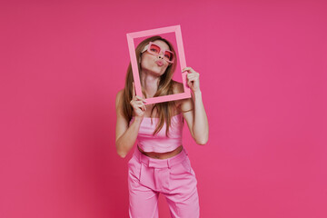 Playful young woman sending a kiss through a picture frame while standing against pink background