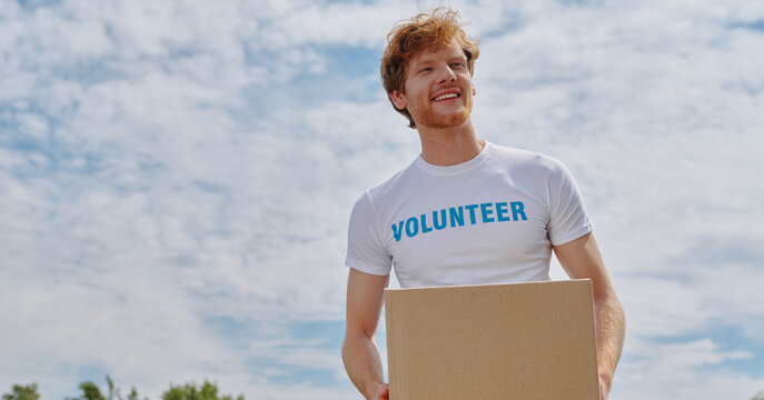 Cheerful Red Hair Man In Volunteer Shirt Carrying Box And Smiling While Standing Outdoors