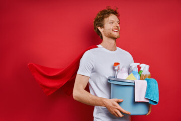 Cheerful man with superhero cape holding bucket with cleaning products against red background