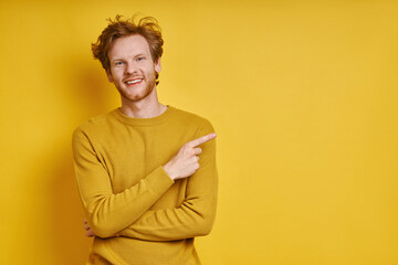Handsome redhead man pointing away and smiling while standing against yellow background