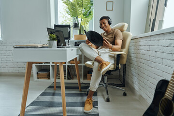 Happy multiracial man in headphones examining vinyl record while sitting at working place