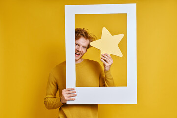 Handsome man holding star while looking through a picture frame against yellow background