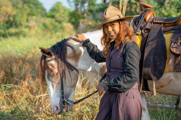 Pretty young Asian woman with cowboy costume smile and stand in front of white horse and also look at camera in field.
