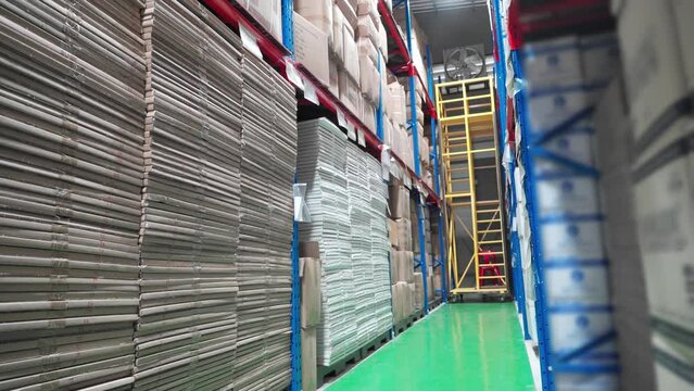 Low angle view of Caucasian male warehouse worker in safety vest, helmet, and face mask to protect Covid-19 holding a cardboard box walking through the aisle  between products on shelves in warehouse