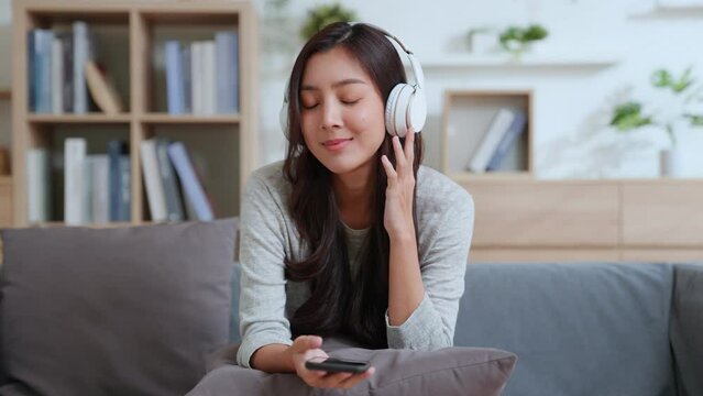 Positive Asian happy woman uses mobile phone and wears bluetooth headphones on couch. smile and feel good. Female listening MP3 music or watching a MV video on smartphone in the living room at home.