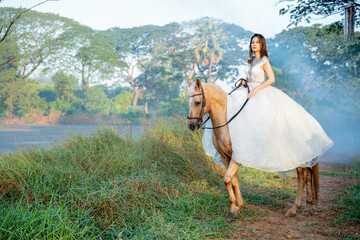 Beautiful woman with white dress stay on brown horse on the road way with background of fog or...