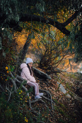 Naklejka premium Young Caucasian girl sitting on tree roots in forest, smiling, looking at camera near river with willow trees in background, wearing hat, pink jacket, sports trousers, sneakers. Autumn walk in forest.