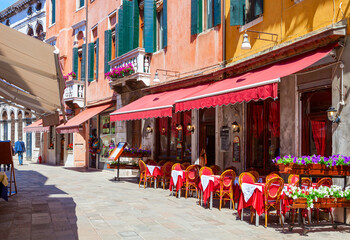 Old street with colorful facades and red  tables of cafe at a sunny morning, Venice, Italy.