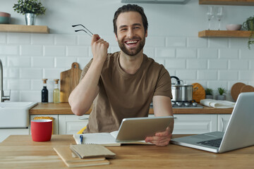 Happy young man using technologies while working at the kitchen island at home