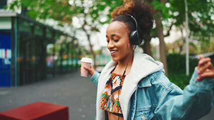 Smiling African girl with ponytail wearing denim jacket, in crop top with national pattern walking down the street listening to music on headphones and dancing