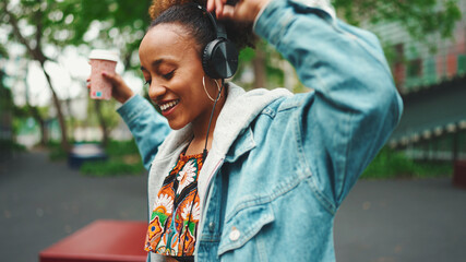 Smiling African girl with ponytail wearing denim jacket, in crop top with national pattern walking down the street listening to music on headphones and dancing
