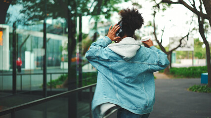 Smiling African girl with ponytail wearing denim jacket, in crop top with national pattern walking down the street listening to music on headphones and dancing