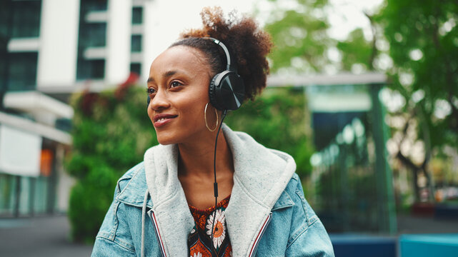 Close-up Cute African Girl With Ponytail Wearing Denim Jacket, In Crop Top With National Pattern, Listening To Music With Headphones In Modern Building Background.