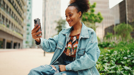Closeup portret cute African girl with ponytail, wearing denim jacket makes stream, video call on smartphone in modern city background. © Andrii Nekrasov