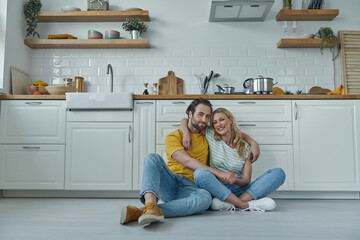 Beautiful young couple embracing while sitting on the floor and leaning at kitchen counter