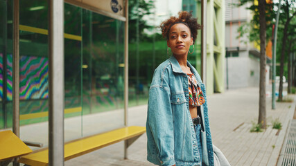 Cute African girl with ponytail, wearing denim jacket, in crop top with national pattern, standing at the bus stop and smiling