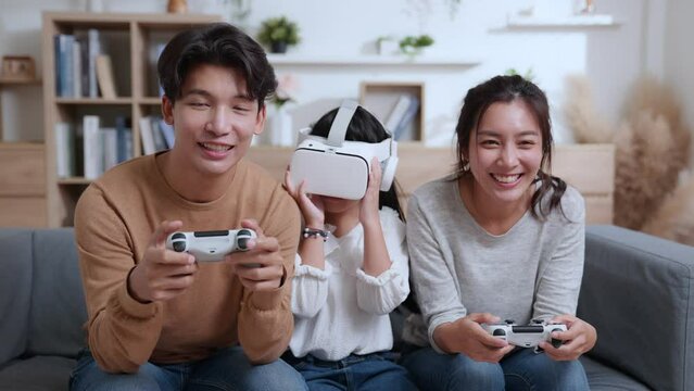 Father and mother playing video game with joystick. A young daughter wearing VR glasses on couch at home. Asian family spending time together on holiday in living room. Attractive happy parents.