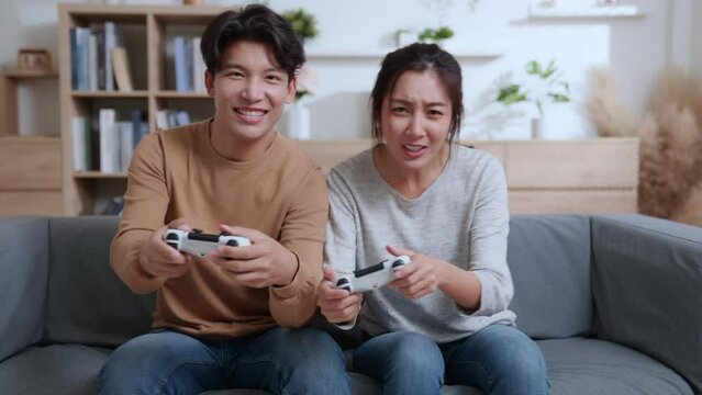 Happy couple asian sitting on the couch playing video games, using joystick controllers. Woman and man in love have fun playing in video games console at home together.