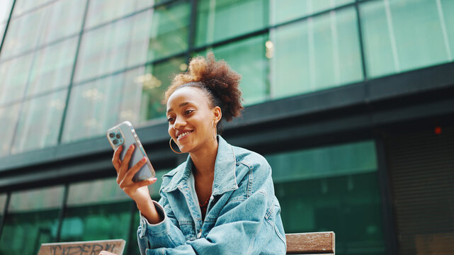 Cute African girl with ponytail, wearing denim jacket, in crop top with national pattern, sitting on bench and using smartphone on modern buildings background.