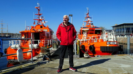 aktiver Senior steht vor orangen Lotsenbooten am Hafen von Travemünde unter blauem Himmel