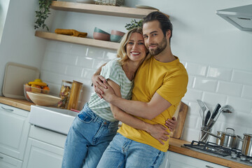 Beautiful young couple embracing and looking at camera while standing at the kitchen
