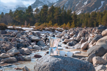 Moka coffee pot and two porcelain cup on big stone against backdrop of mountain river