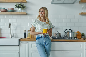 Cheerful young woman holding coffee cup and leaning at the kitchen desk