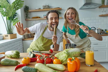 Playful young couple singing and looking at camera while cooking together at the kitchen