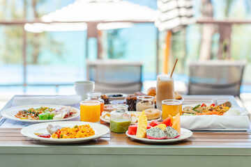 Delicious breakfast set on wooden table next to the pool with sea view. Morning food in tropical modern resort. Assortment of fresh food - fruits, vegan juices, matcha green tea and pastry
