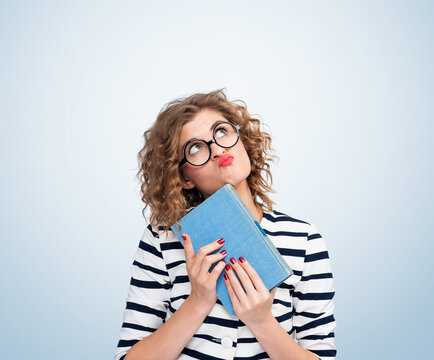 Portrait of a young dreaming girl with glasses holding an old book in her hands and looking up thoughtfully, on light blue background.