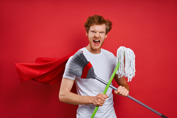 Furious redhead man in superhero cape carrying cleaning equipment while against red background