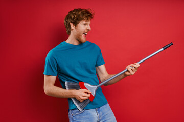 Playful young redhead man using broom like guitar while against red background