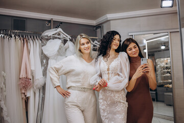 Women taking photographs of a female friend trying on wedding dress. Women in wedding dress fitting room.