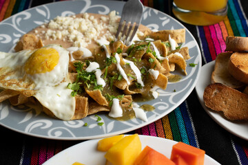 traditional mexican food called chilaquiles in green sauce, with beans, slices of bolillo, tropical fruit of the region: mango, papaya and orange juice, on a typical mexican cloth tablecloth.