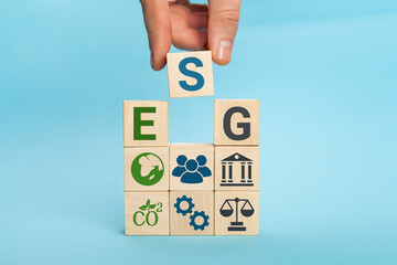 ESG concept of environmental, social and governance. Words ESG on a wood cubes. Hand holds wooden cube with abbreviation ESG standing with other icons on blue background