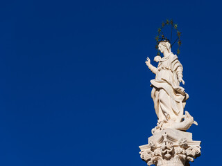 Fototapeta na wymiar Statue of Virgin Mary of the Stars crushing dragon and crescent at the top of an ancient column in the historic center of Lucca, erected in 1687 (with blue sky and copy space)