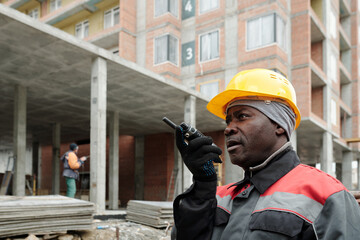 Aged builder in hardhat and workwear speaking in walkie-talkie while working at construction site against unfinished concrete structure