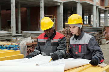 Confident mature foreman in uniform explaining sketch to young female subordinate while pointing at blueprint with architectural plan