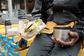 Hand of African American worker of construction site holding cup with hot tea and sandwich while...