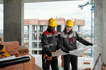 Two intercultural engineers or builders in workwear and hardhats discussing architectural plan or...